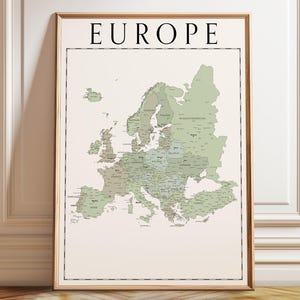 May include: A framed map of Europe, presented in a light brown wooden frame. The map features a beige backdrop with countries rendered in shades of green and grey. Country names are labelled in black. The word "EUROPE" is printed in black at the top.