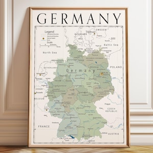 May include: A framed wall map of Germany featuring a sage green landmass against a cream background. The map displays major cities, state borders, and surrounding countries with black text labels. The title GERMANY is printed in large black font.