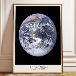 Può includere: Una stampa da parete rettangolare incorniciata che presenta l'iconica fotografia della Terra, la Blue Marble, su uno sfondo nero. Sotto l'immagine, il testo recita: The Blue Marble - Apollo 17, 1972 - in un carattere serif nero e chiaro.
