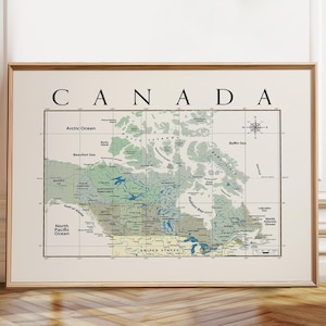 May include: A framed wall art print featuring a detailed map of Canada with the word CANADA printed in large black capital letters above the map. The map displays geographical regions, bodies of water, and a compass rose in a muted color palette.
