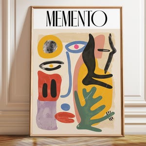May include: A rectangular wall art print in a wooden frame featuring the word MEMENTO in black capital letters at the top. The artwork displays an abstract composition of colorful shapes including yellow, red, green, black, and purple forms on a beige background.