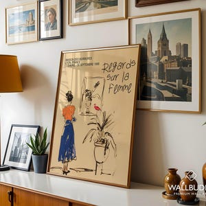 Op de afbeelding: Een poster met een gouden lijst met een zwart-witte illustratie van een vrouw in een blauwe jurk die voor een raam staat. De poster heeft de tekst "Regards sur la Femme" en "Maison des Murmures Lyon France Avril-30 Septembre 1998".