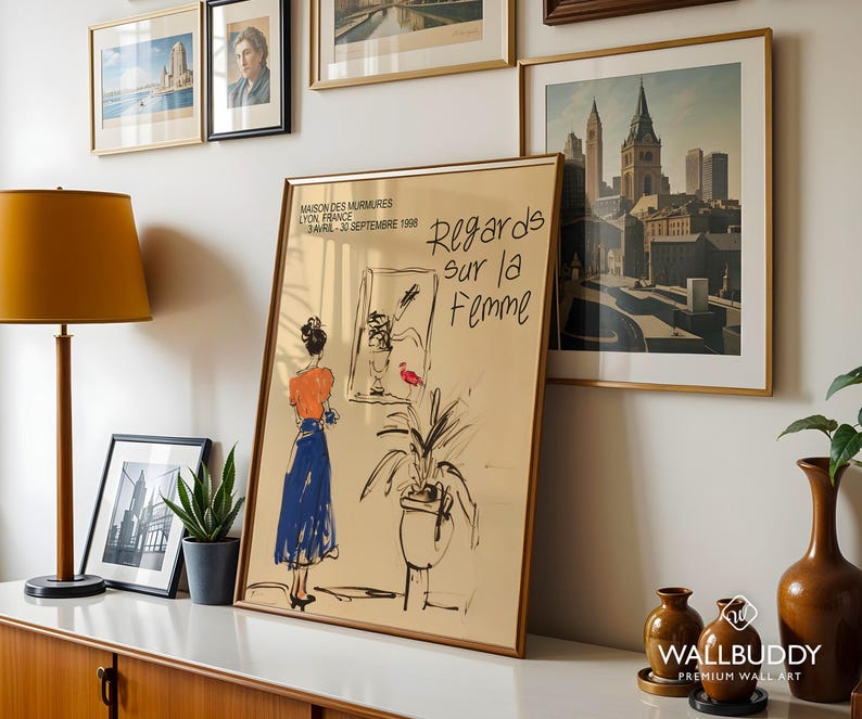 Op de afbeelding: Een poster met een gouden lijst met een zwart-witte illustratie van een vrouw in een blauwe jurk die voor een raam staat. De poster heeft de tekst "Regards sur la Femme" en "Maison des Murmures Lyon France Avril-30 Septembre 1998".
