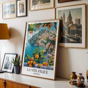 Peut inclure: Une impression d'art verticale encadr&eacute;e intitul&eacute;e LA VITA FELICE Amalfi Coast, pr&eacute;sentant une illustration vibrante de la c&ocirc;te italienne avec une eau turquoise, des b&acirc;timents color&eacute;s &agrave; flanc de colline et des branches charg&eacute;es d'agrumes orange.