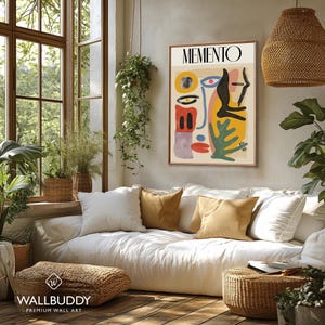May include: A framed wall art print titled MEMENTO featuring an abstract composition of organic shapes in yellow, black, red, blue, and green on a beige background. The artwork displays stylized facial features and geometric forms in a vertical layout.