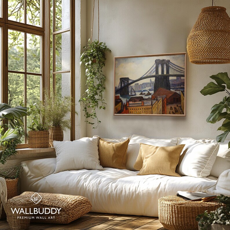 May include: A framed wall art print depicting the Brooklyn Bridge in a painterly style with muted blue, brown, and orange tones. The artwork is displayed on a light-colored wall above a white sofa with tan throw pillows in a sunlit room.
