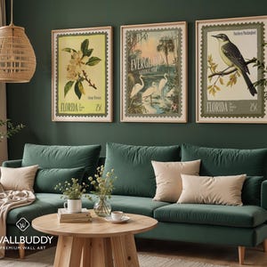 May include: Three framed vintage-style prints depicting Florida scenes. The prints feature a magnolia flower, Everglades National Park with herons, and a Northern Mockingbird. The prints are displayed above a green velvet sofa with beige cushions.