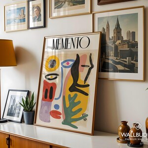 May include: A framed vertical poster titled MEMENTO featuring an abstract composition of organic shapes in yellow, red, blue, green, and black. The artwork displays a stylized face with a prominent eye, a red mouth, and various geometric forms.