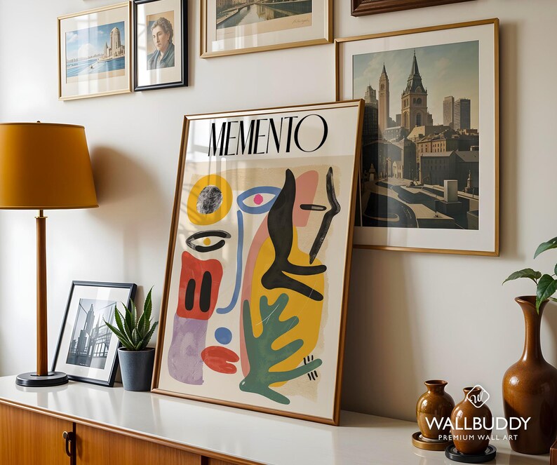 May include: A framed vertical poster titled MEMENTO featuring an abstract composition of organic shapes in yellow, red, blue, green, and black. The artwork displays a stylized face with a prominent eye, a red mouth, and various geometric forms.