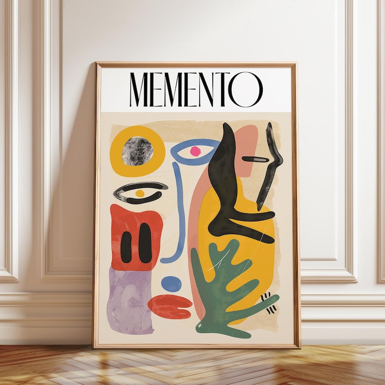 May include: A rectangular wall art print in a wooden frame featuring the word MEMENTO in black capital letters at the top. The artwork displays an abstract composition of colorful shapes including yellow, red, green, black, and purple forms on a beige background.