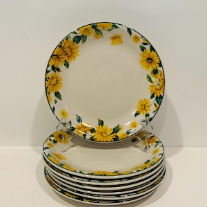 May include: A stack of white ceramic plates with a yellow sunflower design around the rim. The plates have a dark green border and are displayed on a clear plastic stand. The sunflower design includes yellow flowers and green leaves.