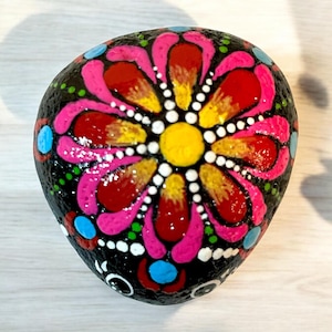 Op de afbeelding: Een handgeschilderde steen met een zwarte achtergrond en een bloemenontwerp in roze, rood, geel en wit.