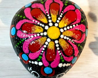 Puede incluir: Una piedra pintada a mano con fondo negro y un diseño de flor rosa, roja, amarilla y blanca.