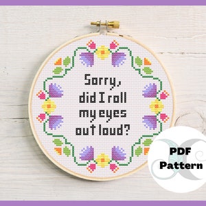May include: A cross-stitch pattern featuring a floral wreath with the text "Sorry, did I roll my eyes out loud?" in black text. The pattern is in a wooden embroidery hoop and is labeled "PDF Pattern".