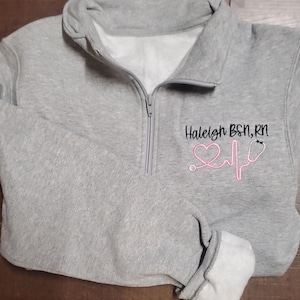 May include: Gray fleece pullover with a zipper and a pink heart and stethoscope graphic. The text "Haleigh BSN, RN" is embroidered on the front.