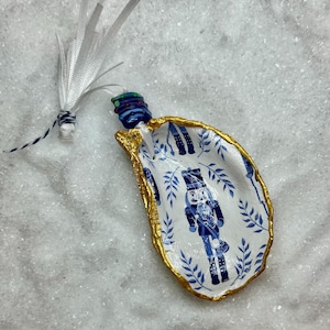 May include: A decorative oyster shell trinket dish with a gold rim, featuring a blue and white nutcracker design. The shell has a blue and white ribbon attached for hanging. The dish is on a white, textured surface.