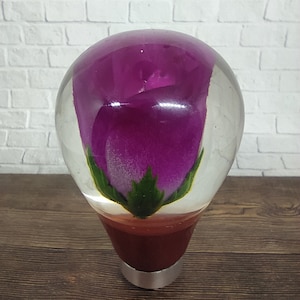 Pode incluir: Uma alavanca de câmbio de vidro transparente com uma rosa roxa dentro. A rosa tem folhas verdes e está encapsulada em resina.