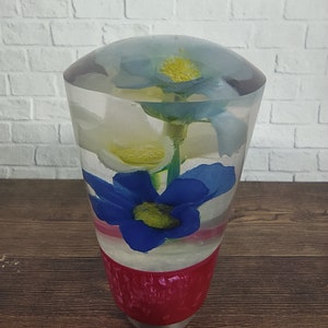 Könnte beinhalten: Ein transparenter Glasschaltknauf mit roter Basis und drei Blumen im Inneren. Die Blumen sind blau, weiß und gelb.