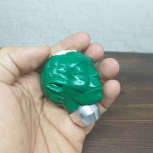 May include: Green and white figurine of a head with a silver base. The head has a closed-eye expression and a pointed ear.