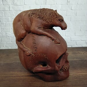 Peut inclure: Une sculpture en terre cuite d'un crâne sur lequel repose un animal stylisé, peut-être un chien. La sculpture est de couleur brune et a une surface texturée.