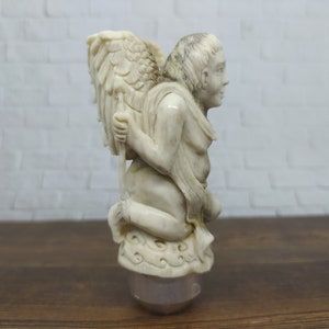 Può includere: Una statuetta bianca intagliata di un angelo con le ali, inginocchiato e che tiene un bastone. L'angelo indossa una veste fluida e ha un viso dettagliato.
