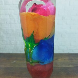 Peut inclure: Un cylindre en verre transparent contenant une rose rouge, orange, rose et bleue. Les roses sont superposées et enchâssées dans de la résine transparente.