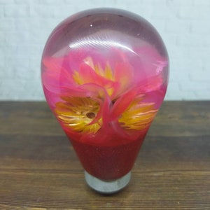 Op de afbeelding: Een pookknop van helder hars met een roze en gele bloem erin. De knop is rood aan de onderkant en heeft een zilveren basis.