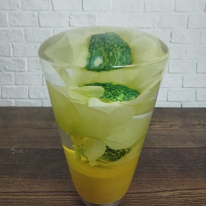 Pode incluir: Um vaso de vidro transparente com base amarela cheio de uma flor branca e folhas verdes. A flor e as folhas são preservadas em resina.