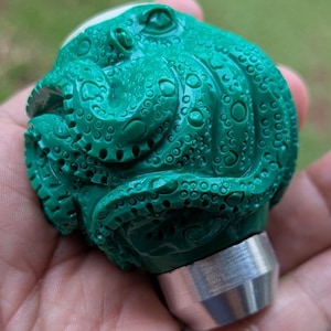 May include: A detailed, green octopus sculpture with a textured surface. The sculpture has a silver metal base. The octopus design features intricate details, including suckers and a rounded head. The sculpture is a unique decorative item.