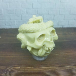 May include: Ivory-colored skull sculpture with a snake coiled around it. The skull has detailed features, including eye sockets, teeth, and a textured surface. The snake is also intricately carved, adding to the sculpture's gothic aesthetic.