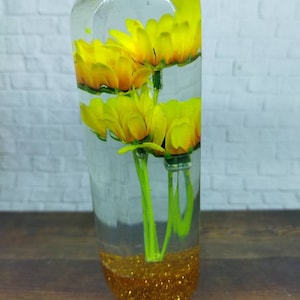 May include: A clear glass cylinder filled with water and yellow flowers. The bottom of the cylinder is filled with gold glitter.
