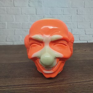Puede incluir: Una cara de payaso de plástico naranja y blanco brillante con una amplia sonrisa y los ojos cerrados.