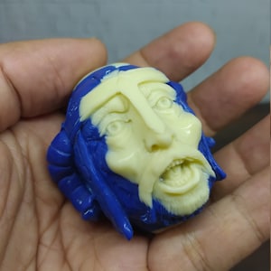 Puede incluir: Una escultura detallada de una cara con cabello azul y una cara, barba y tocado en forma de cruz de color crema. La cara tiene la boca abierta y parece angustiada. La escultura se sostiene en una mano.