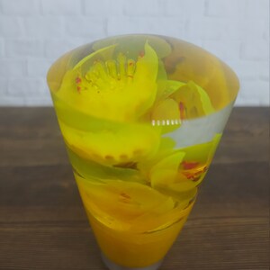 May include: A clear glass paperweight with yellow flowers encased inside. The flowers are arranged in a circular pattern and have a translucent appearance.