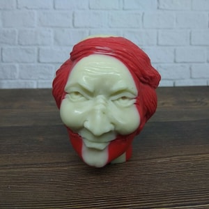 Puede incluir: Una figura vintage y novedosa de una cara con cabello y acentos rojos. La cara es de color pálido con detalles rojos alrededor de la boca y el cabello. La figura tiene una expresión severa.