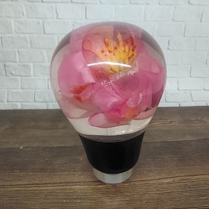 Pode incluir: Uma alavanca de câmbio de vidro transparente com uma flor rosa encapsulada dentro. A flor tem detalhes amarelos e vermelhos.