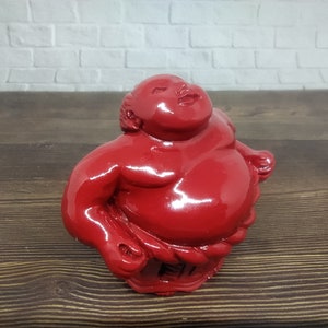 May include: A red, glossy, ceramic figurine of a sumo wrestler. The figurine is standing with its arms raised.