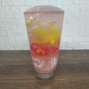 May include: A clear glass shift knob with a pink, yellow, and red floral design inside. The knob is on a wooden surface.