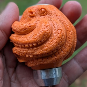 Op de afbeelding: Een oranje, ingewikkeld gebeeldhouwd object in de vorm van een octopus met een getextureerd oppervlak en een zilvermetalen basis. Het octopusontwerp heeft gedetailleerde ogen en tentakels, waardoor een uniek en artistiek stuk ontstaat.