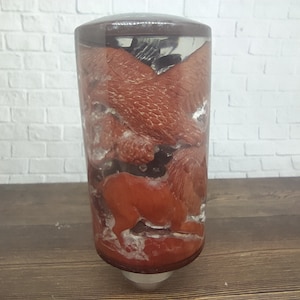 Op de afbeelding: Een doorzichtige glazen cilinder met een bruine bovenkant bevat een rood en wit dierlijk beeldje. Het beeldje is gedeeltelijk ondergedompeld in een heldere vloeistof.