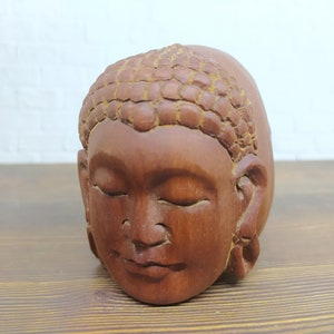 May include: A wooden sculpture of a Buddha head with closed eyes. The sculpture is carved with intricate details and has a reddish-brown color.