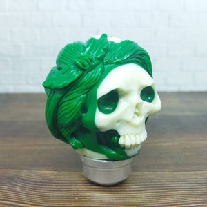 Puede incluir: Una figura decorativa de calavera con un diseño de hojas verdes alrededor de la cabeza y una cara de calavera blanca. La figura se asienta sobre una base plateada. La calavera tiene cuencas de ojos verde oscuro y una mandíbula detallada.