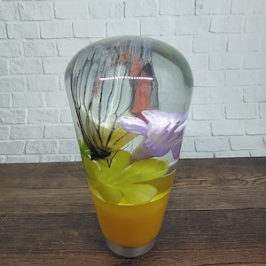 Pode incluir: Pomo de mudança decorativo em resina com asa de borboleta e flor roxa. A parte inferior é amarela sólida. O pomo está sobre uma parede de tijolos brancos e uma superfície de madeira.