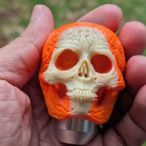 May include: A detailed, orange and off-white skull sculpture with a silver base. The skull features intricate carvings and a realistic design. The eye sockets and nasal cavity are dark orange, contrasting with the off-white skull.