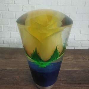 Pode incluir: Um peso de papel de vidro transparente com uma rosa amarela dentro. A rosa é cercada por folhas verdes e água azul. O peso de papel está sobre uma superfície de madeira.