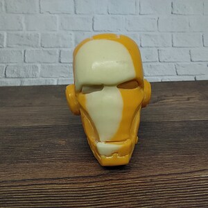 Puede incluir: Una escultura de cabeza impresa en 3D con un esquema de pintura amarillo y blanco. La cabeza tiene una forma humanoide con una cara plana y ancha y una nariz prominente.