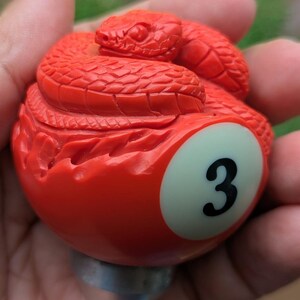 Puede incluir: Una bola de billar roja brillante con el número 3, coronada con una escultura detallada de una serpiente enrollada. La serpiente está intrincadamente tallada, agregando textura al diseño. La bola tiene un acabado liso y una base de metal.