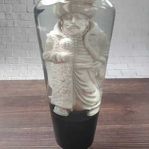 Könnte beinhalten: Eine weiße Figur eines Mannes mit Turban und traditioneller Kleidung ist in einer durchsichtigen Glaskugel eingeschlossen. Die Kugel ist auf einem schwarz-weißen Sockel montiert.