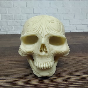 Peut inclure: Un crâne en céramique blanche avec des sculptures complexes sur le front et autour des orbites. Le crâne est posé sur une surface en bois.