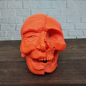 Puede incluir: Una decoración de Halloween de color naranja brillante que representa un cráneo con una cara humana en un lado. El cráneo está detallado con dientes y cuencas de los ojos, mientras que la cara humana tiene rasgos realistas.